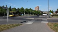 En gul buss kör på en bred väg vid en korsning med trafikskyltar och övergångsställe, omgiven av grönska och byggnader i bakgrunden.