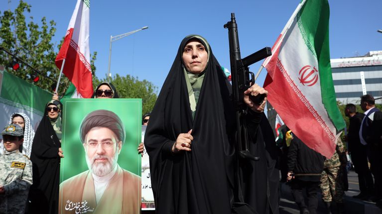 Iranilaisnainen pitelee asetta mielenosoituksessa, vieressä Iranin lippuja ja ajatollah Mojtaba Khamenein valokuva.