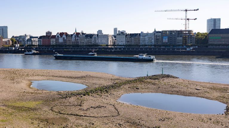 Två stora vattenpölar på en torr flodstrand med en pråm som passerar på floden och stadshus i bakgrunden.