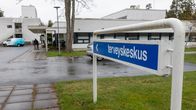Terveyskeskus-kyltti. Taustalla valkoinen terveyskeskus rakennus. Henkilö työntää pyörätuolia.