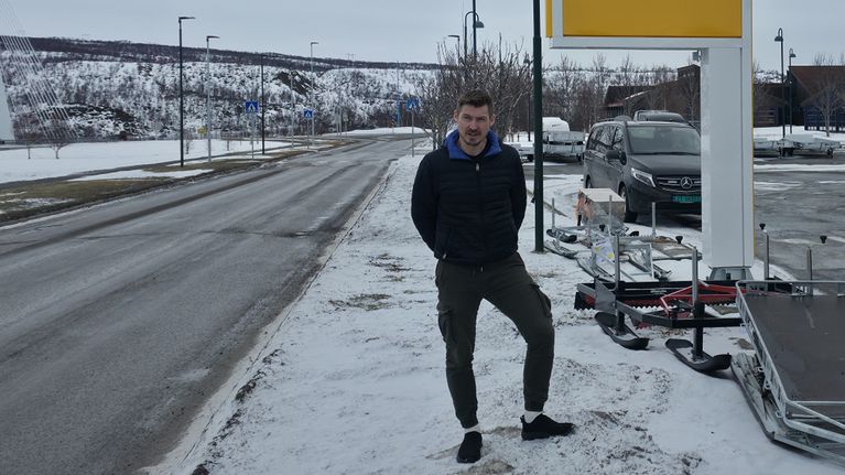 Tummiin vaatteisiin pukeutunut mies seisoo huoltoaseman hintakyltin edessä. 