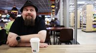 Tommi Ruotsalainen istuu liikennemyymälän pöydän ääressä, edessään kahvikuppi. Hänellä on musta paita ja hattu, ja ranteessaan älykello. Taustalla näkyy kahvilan sisustus ja asiakkaita.