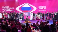 Ett tjugotal leende människor står på scen och vinkar. I mitten av bilden syns den ryska flaggan och texten "Intervision Russia '25".