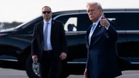 Trump näyttää peukaloa kameralle. Taustalla suuri musta auto ja henkivartija.