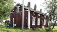 Ett gammalt rödmålad trähus med vita knutar i lantlig miljö omgiven av grönska och träd.