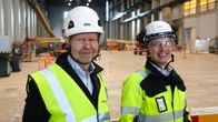 Två personer med skyddshjälmar och reflexvästar står i en stor industrilokal.