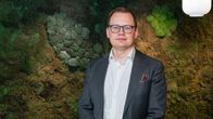 En man i vit skjorta, mörk kavaj och glasögon ser in i kameran. 