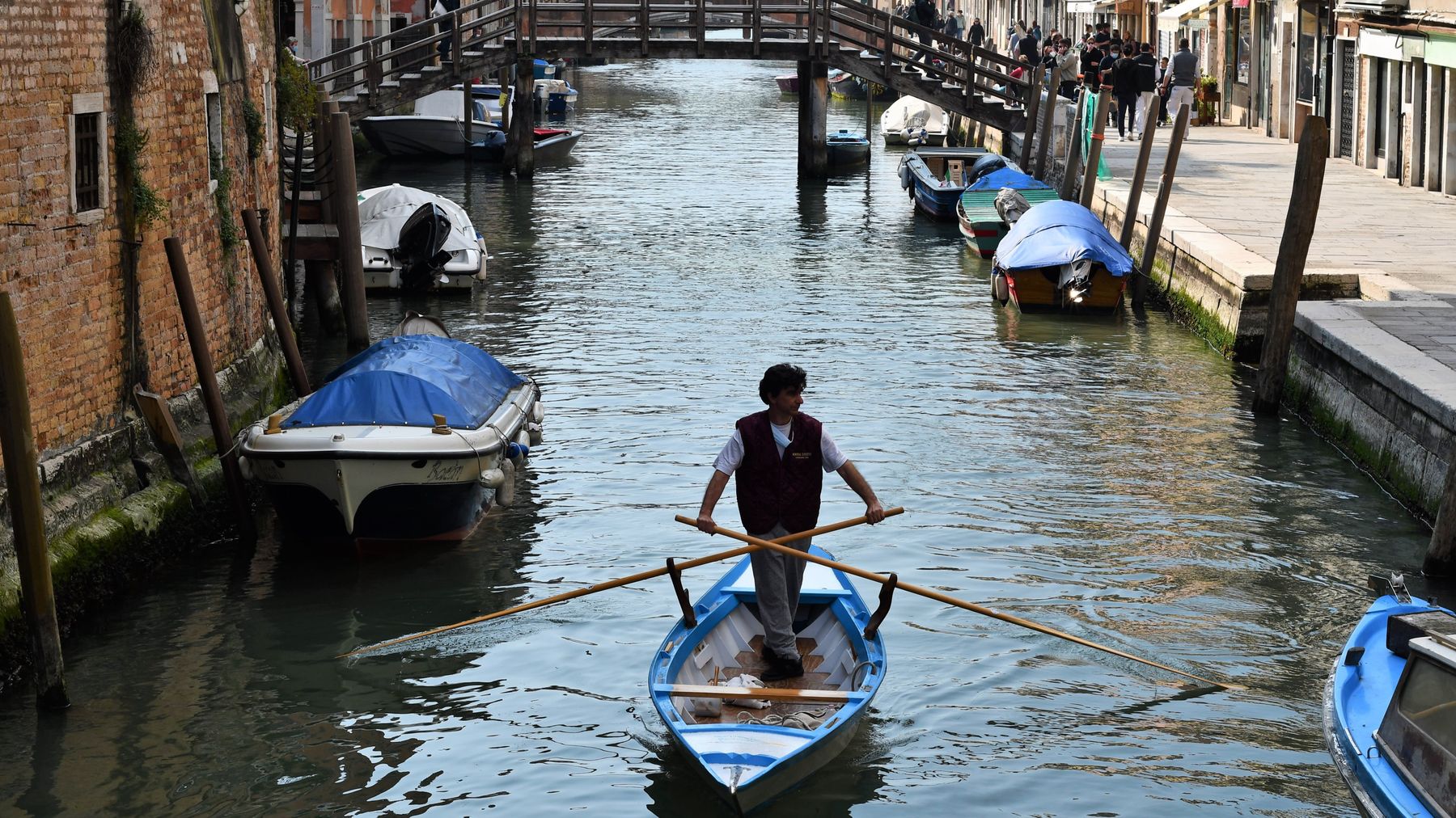 En man står i en speciell roddbåt som används i kanalerna i Venedig. Längs kanalens kant ligger båtar förtöjda med pressningar på och det är tomt på gatan invid kanalen.