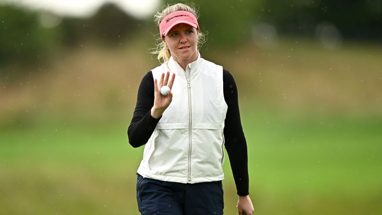 Henkilö seisoo nurmikentällä golfmaila kädessä, pitää golfpalloa oikeassa kädessään ja katsoo kameraan.