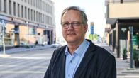 En person i kostym står på en tom gata i en stadsmiljö med butiker och kontorsbyggnader i bakgrunden.
