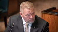 Bilden visar en man i kostym och slips, sittande vid ett bord med en mikrofon framför sig.