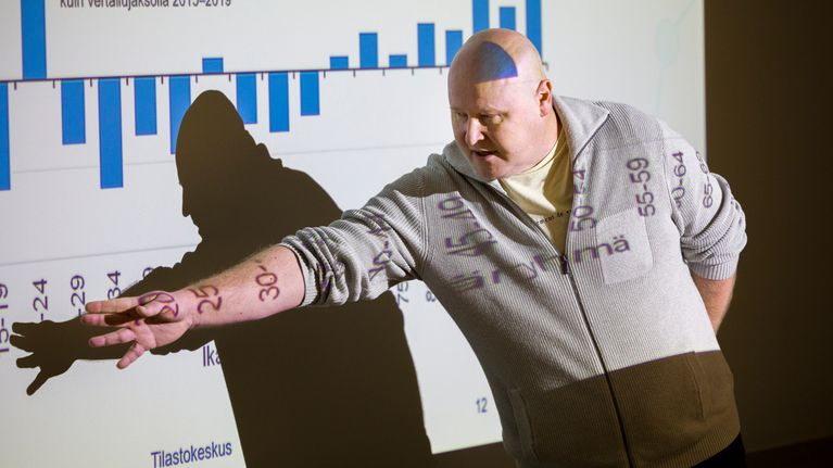 A bald-headed man points to figures on a screen in a dark room, with numbers projected onto his shirt and arm.