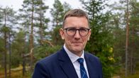 Keski-ikäinen mies seisoo tummassa puvussa ja silmälaseissa metsän keskellä, taustalla havupuita.