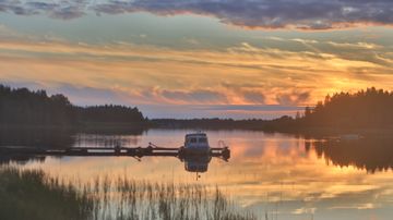 Järvimaisema auringonlaskussa, laiturille kiinnitetty vene ja heijastuksia veden pinnalla, taustalla metsä.