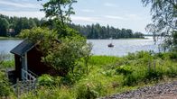 Sommarlandskap med en röd stuga vid stranden och en segelbåt i vattnet.