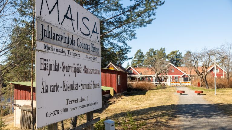 Suuri kyltti: Juhlaravintola Maisa tarjoaa palveluita ja ruokaa luonnonläheisessä ympäristössä Maisansalossa, Tampereella. Taustalla punainen rakennus.