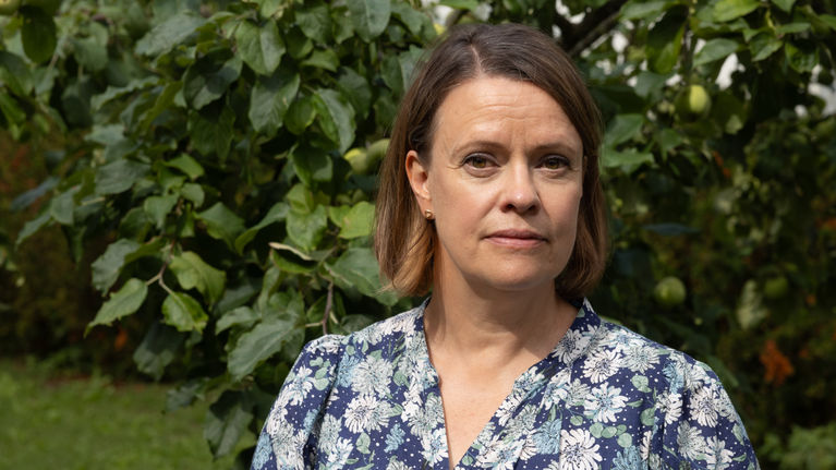 A woman in a blue floral dress stands in a green garden, looking at the camera. 