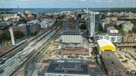 Tampereen keskusta ja rakennuksia rautatieaseman läheisyydessä. Etualalla on rautatie ja junia, ja taustalla näkyvät modernit rakennukset sekä järvi ja vehreitä alueita. Taivas on pilvinen, mutta kirkas.