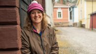 En kvinna sitter lutad mot en trävägg längs en kullerstensgata i ett område med färgglada trähus. Hon är klädd i en brun jacka och rosa mössa.
