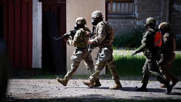 Four soldiers walking wearing heavy gear in an urban setting, first two in khaki uniforms and carrying guns.
