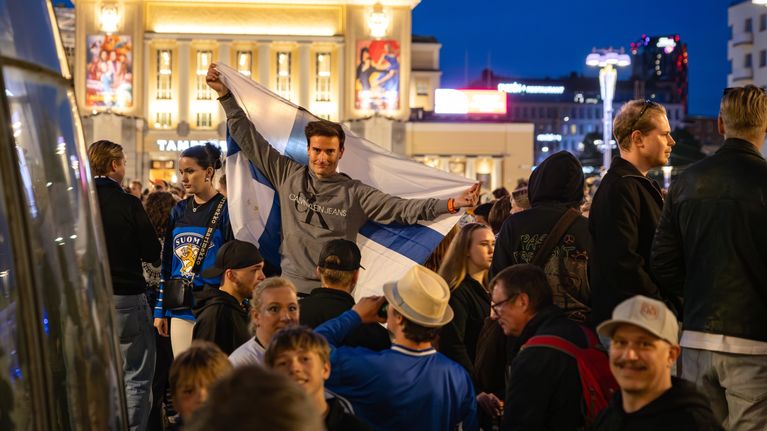 Mies poseeraa suihkulähteen reunalla kameralle. Hän on vetänyt taustakseen Suomen lipun.