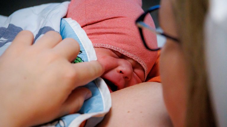 Vuorokauden vanha vauva käärittynä värikkääseen peittoon äidinsä rinnalle. Vauvaa itkettää hiukan.