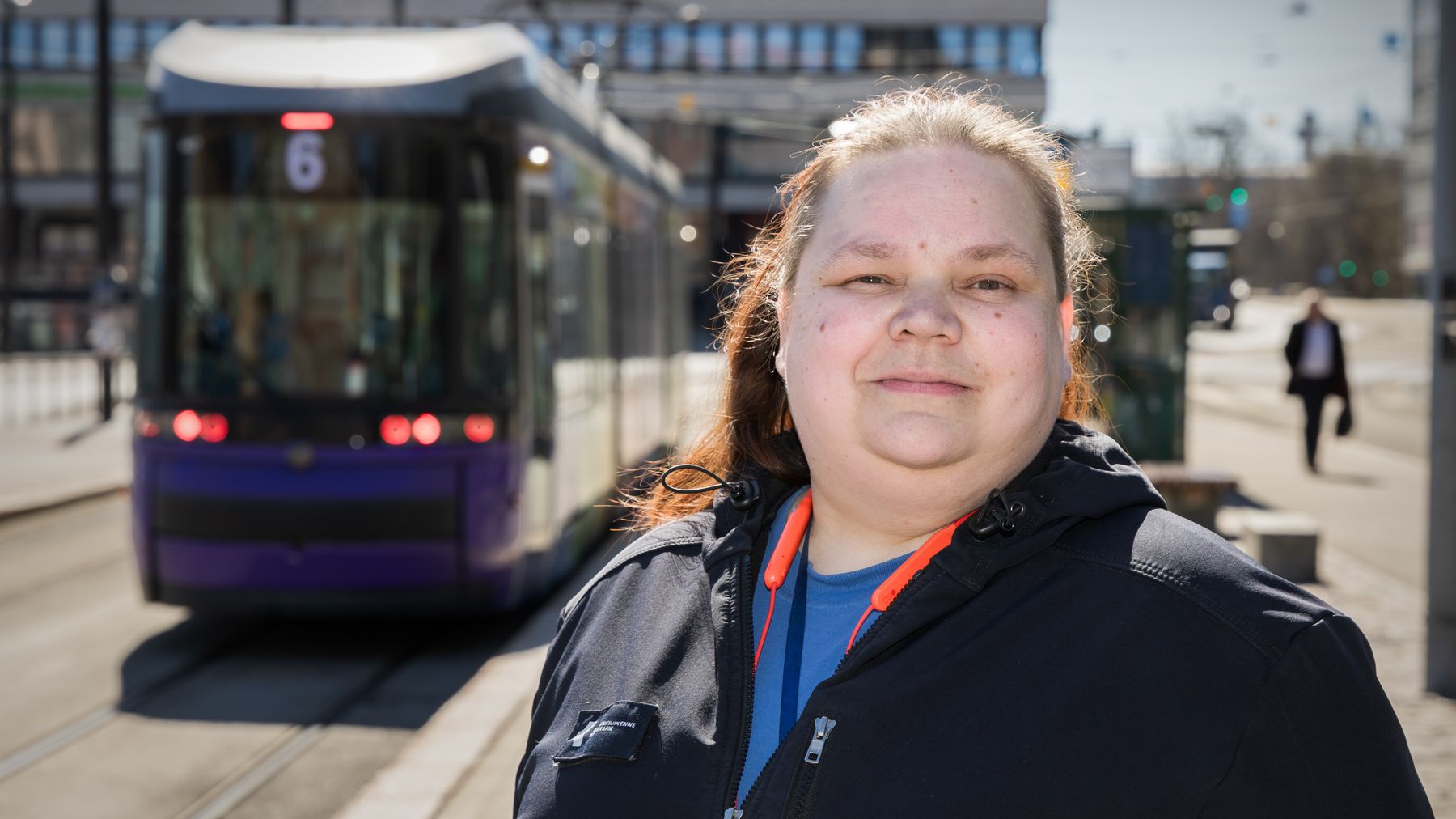 En kvinna står vid en spårvagnshållplats med en spårvagn i bakgrunden.