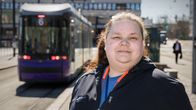 En kvinna står vid en spårvagnshållplats med en spårvagn i bakgrunden.