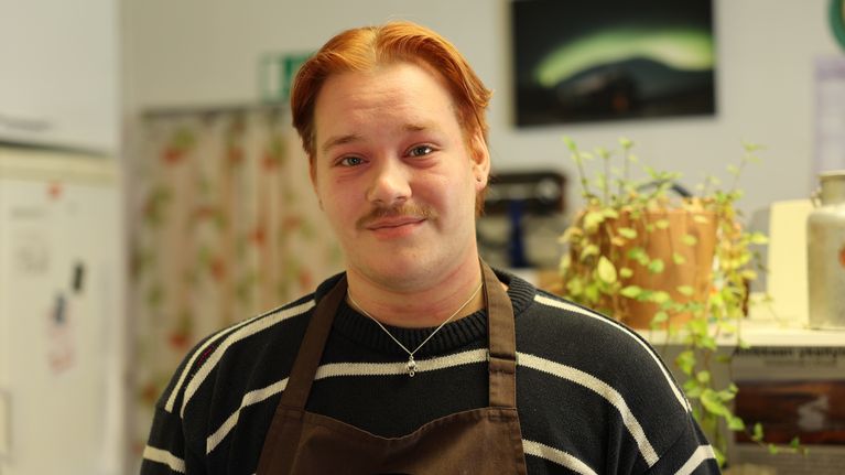 Kuvassa ihminen vienosti hymyilee. Yllään hänellä on essu. Hiukset ovat lyhyet ja oranssihkot.