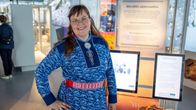 Nainen seisoo sinisessä saamenpuvussa näyttelyn edessä, taustalla vitriinejä ja infotauluja, joissa on tekstiä.