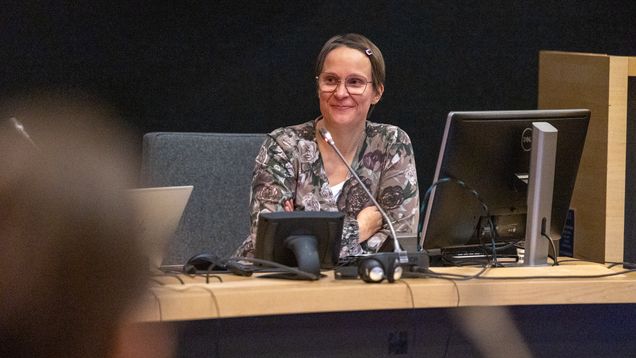 Henkilö istuu neuvottelupöydän ääressä mikrofonin ja tietokoneiden edessä, taustalla tummat seinät.