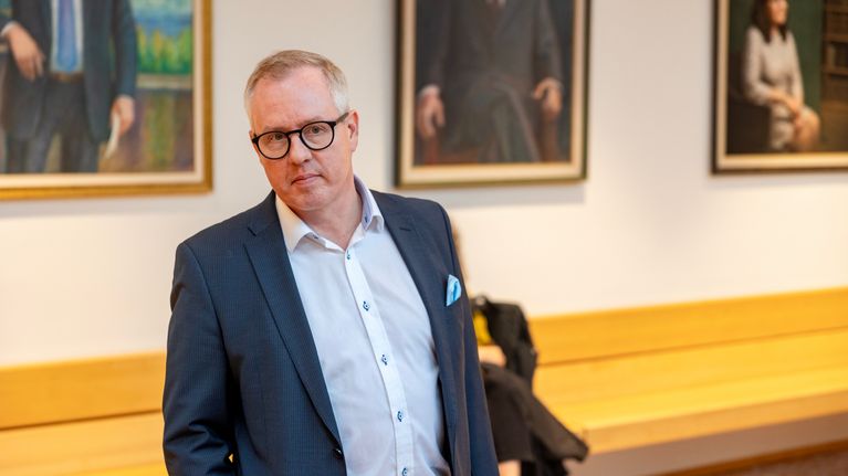 Tumman siniharmaaseen takkiin pukeutunut Vernu Vasunta katsoo eteenpäin kohti kameraa oikeussalissa.