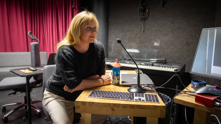 Susa Saukko istuu työtuolilla nojaten puiseen pöytään studiotilassa. Pöydällä on muumitalominiatyyri ja takana viininpunainen molton-verho.