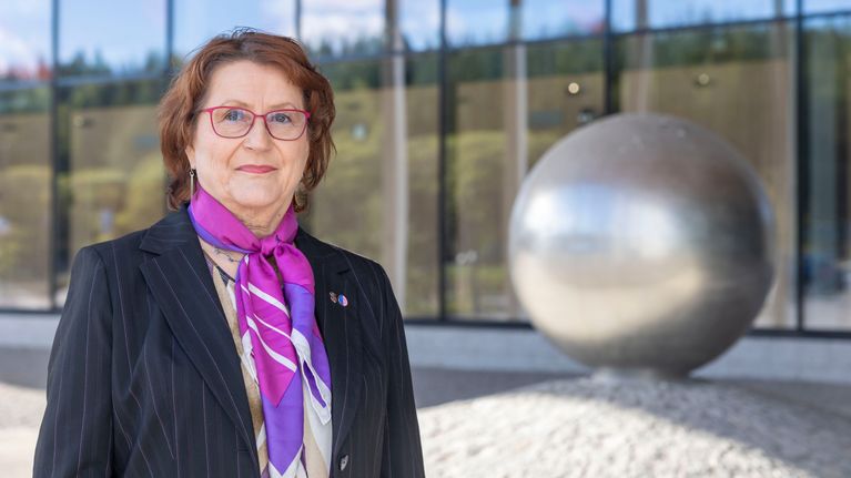 Outi Keinänen taustallaan lasiseinä ja metallinen pallopatsas.