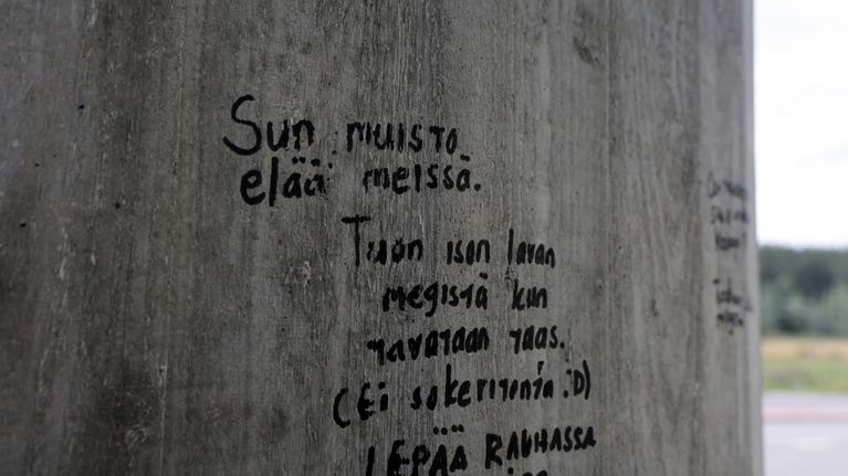 Betonipylvääseen on kirjoitettu mustalla tekstillä viestejä, kuten "Sun muisto elää meissä". Taustalla näkyy metsää.