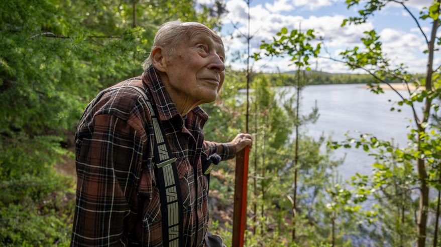 Iäkäs mies seisoo metsän reunassa joen varrella, taustalla vettä ja puita.
