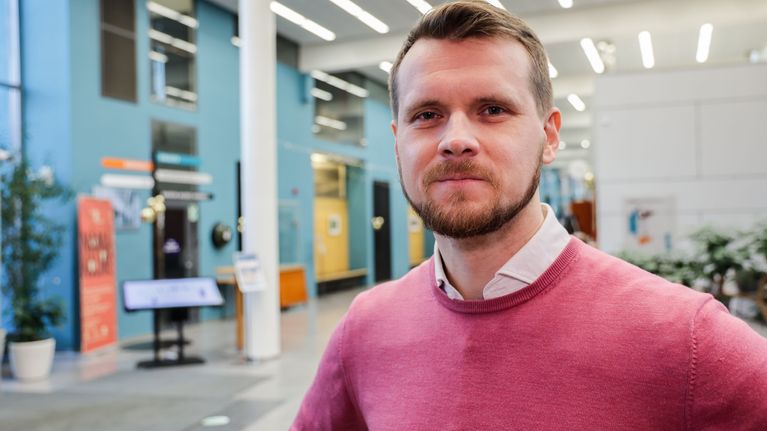Eero Vainio, en person i rosa tröja står inomhus i en ljus och modern byggnad.