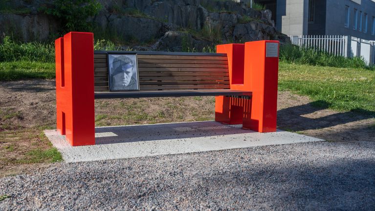 A red-sided park bench in front of a fenced building.
