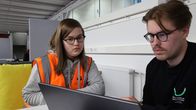 Oranssiin huomioliiviin sonnustautunut nuori naisoletettu kurkistaa läppäriä, jota käyttää tummaan paitaan sonnustautunut nuori miesoletettu. Kummallakin henkilöllä on silmälasit.