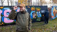 Etualalla mies vihreässä takissa ja takana musta hupparinen henkilö spreijaa luvalliseen graffitiseinään.