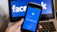 Smarttelefon där inloggning till Facebook visas på skärmen. I bakgrunden en datorskärm där man ser texten Facebook.