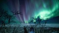Kaksi henkilöä katsoo lumisessa talvimaisemassa taivaalle, jossa näkyy revontulia.