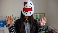 Japanilainen nainen seisoo kotinsa olohuoneessa, heiluttaa käsiään ja hänen naamallaan on naamio, jossa on kuva hänen sarjakuvahahmostaan joka on pyöreähkö, valkoinen hahmo, jolla on punainen kaulaliina ja tonttulakki.