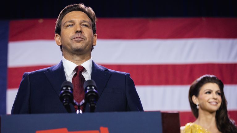 Floridas guvernör Ron DeSantis i röd slips och blå kostym bakom en talarstol fotograferad snett nerifrån. 