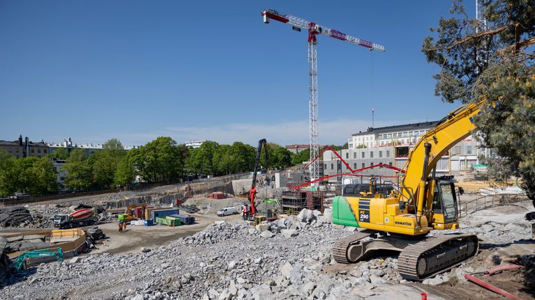 Kivimursketta, käytetyistä autonrenkaista tehtyjä räjäytysmattoja ja koneita työmaakuopassa, jonka takana vihreitä puita ja kerrostaloja.