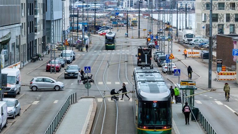 Laajassa kaupunkimaisemassa näkyy raitiovaunuja, lastenvaunujen kanssa suojatietä ylittäviä henkilöitä, autoja ja pyöräilijöitä liikkeessä.