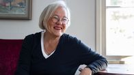 Vanhempi nainen hopeisilla hiuksilla ja mustassa paidassa istuu penkillä pöytään nojaten ja hymyilee aurinkoisesti.