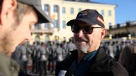 Kaksi miestä keskustelee ulkona, toisella on päässään lippis, ja hän pitää mikrofonia, jossa lukee "yle". Taustalla näkyy sotilaita.