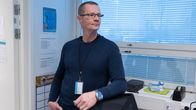 Mies seisoo toimistossa pukeutuneena siniseen neulepaitaan ja farkkuihin. Hänellä on kaulassa henkilökortti ja kädessään kello.