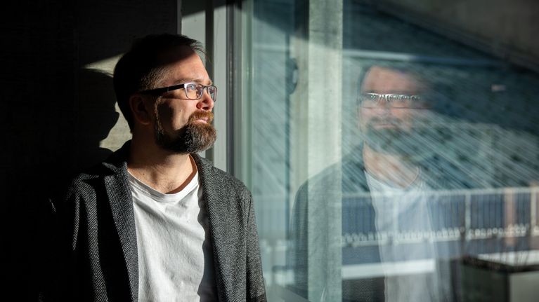 Mies seisoo sisätiloissa, katsellen ikkunasta ulos. Hänen vaatteensa koostuvat tummasta takista ja vaaleasta T-paidasta. Aurinko luo varjostuksia huoneeseen, ja lasipinta heijastaa maisemaa ulkopuolelta.